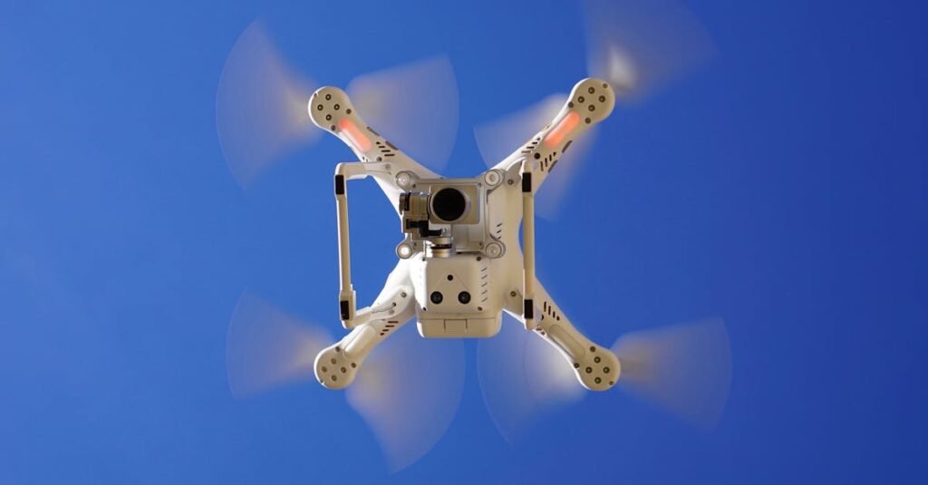 Aerial view of white drone flying with rotating blades against a clear blue sky, capturing video footage.